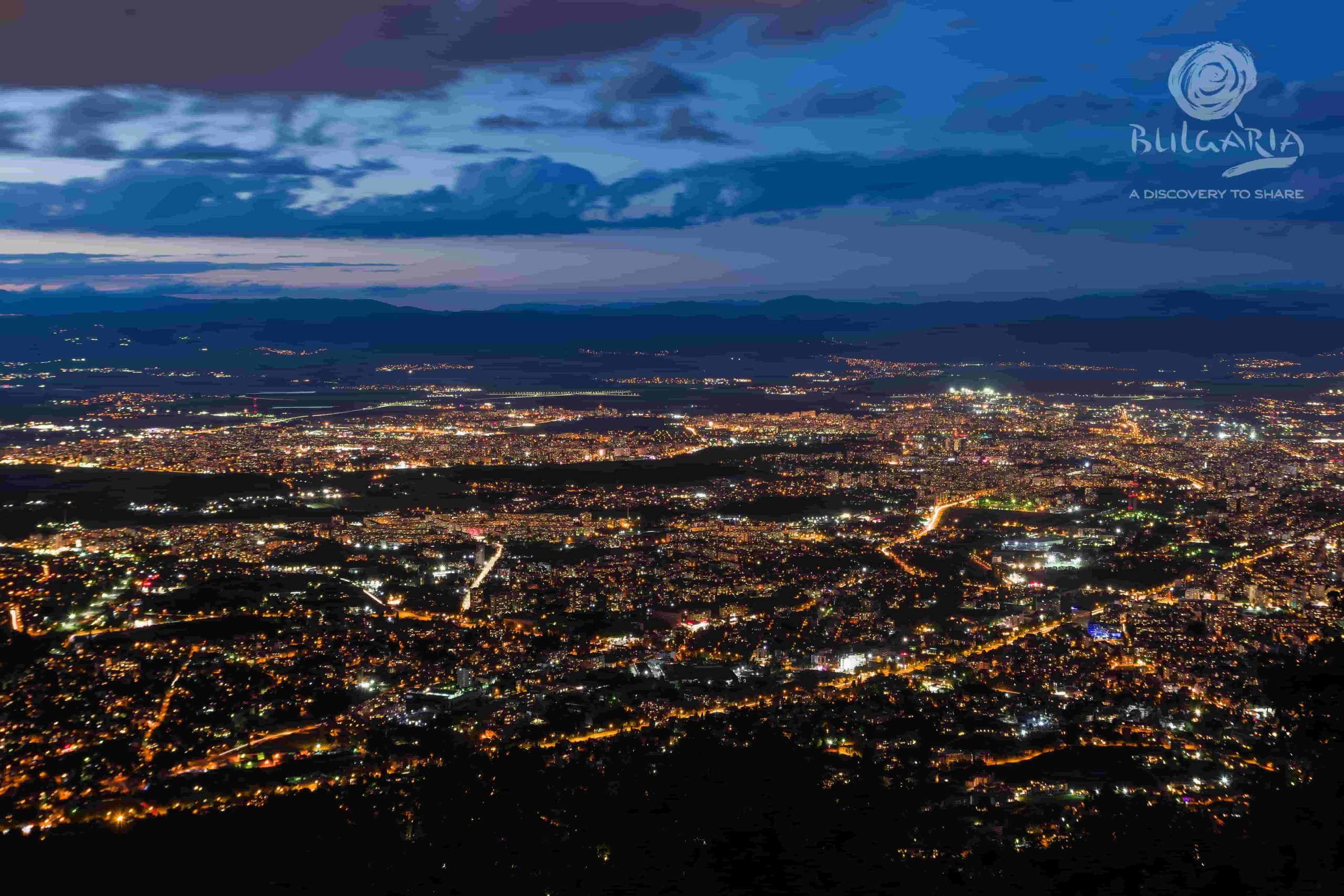 A breathtaking night view of the city, captured from a mountain peak. The city lights twinkle below, creating a mesmerizing sight.