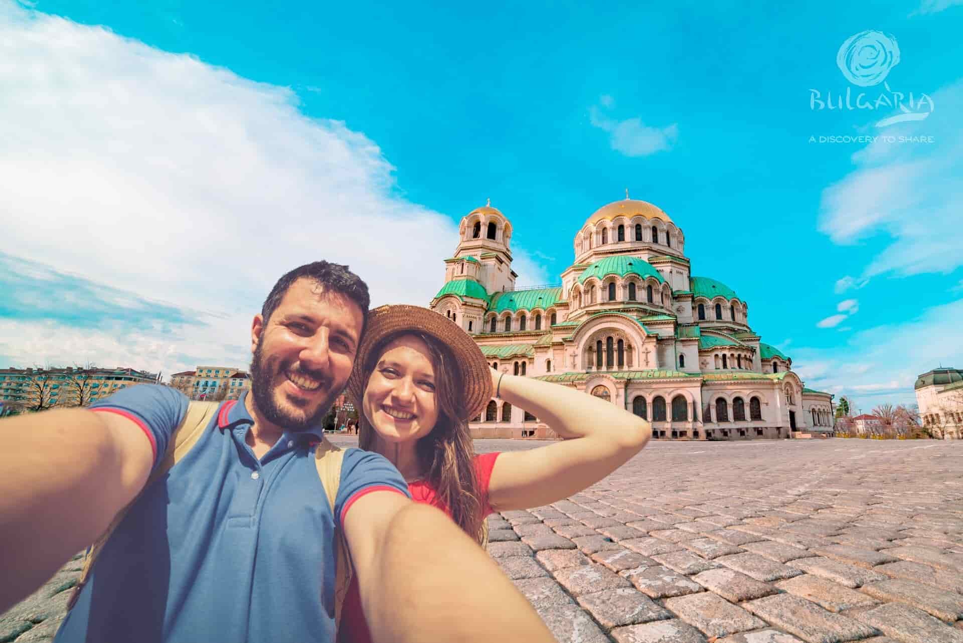 A couple smiling and taking a selfie in front of a beautiful church.
